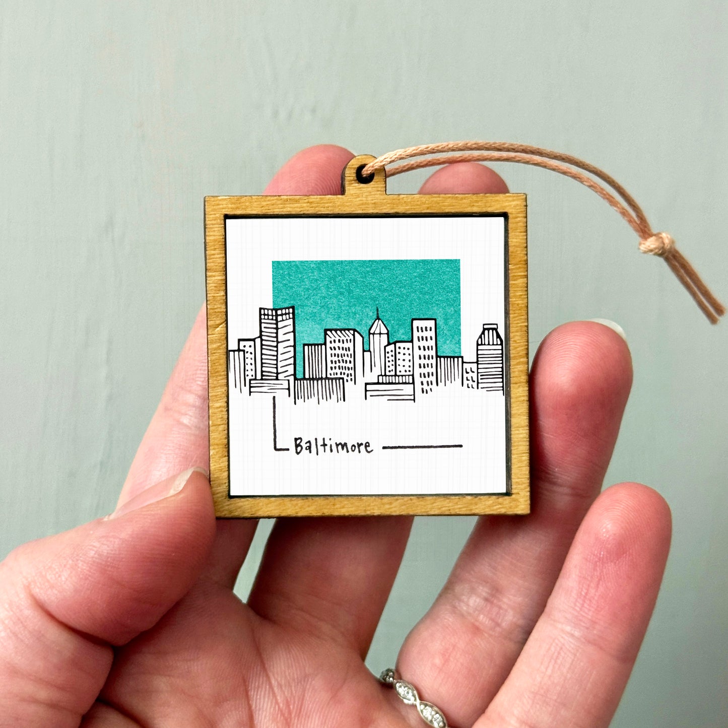 A hand holding a small wooden frame with a drawing of a city skyline and the word "Baltimore" written on it.