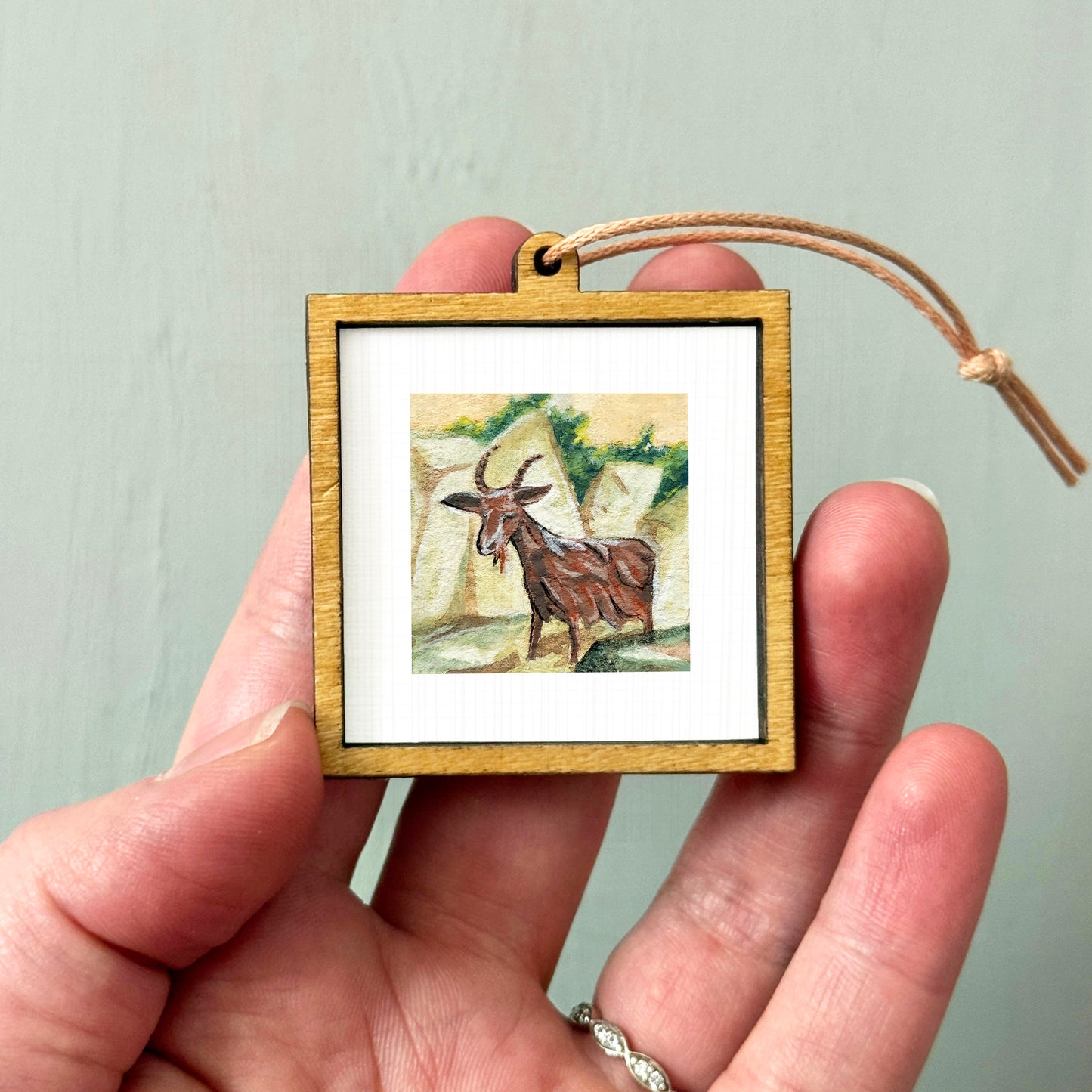 A person's hand holding a small wooden frame containing a painting of a goat.