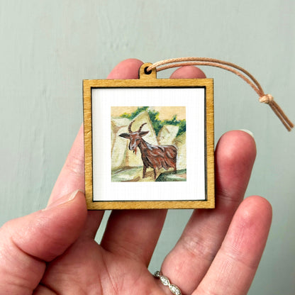 A person's hand holding a small wooden frame containing a painting of a goat.