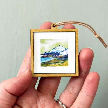 A hand holding a small wooden frame containing a watercolor painting of a mountain landscape.