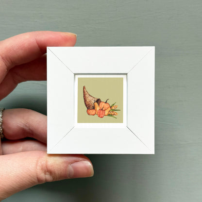 A hand is holding a small white square frame containing a picture of a Thanksgiving turkey and other fall-themed items.