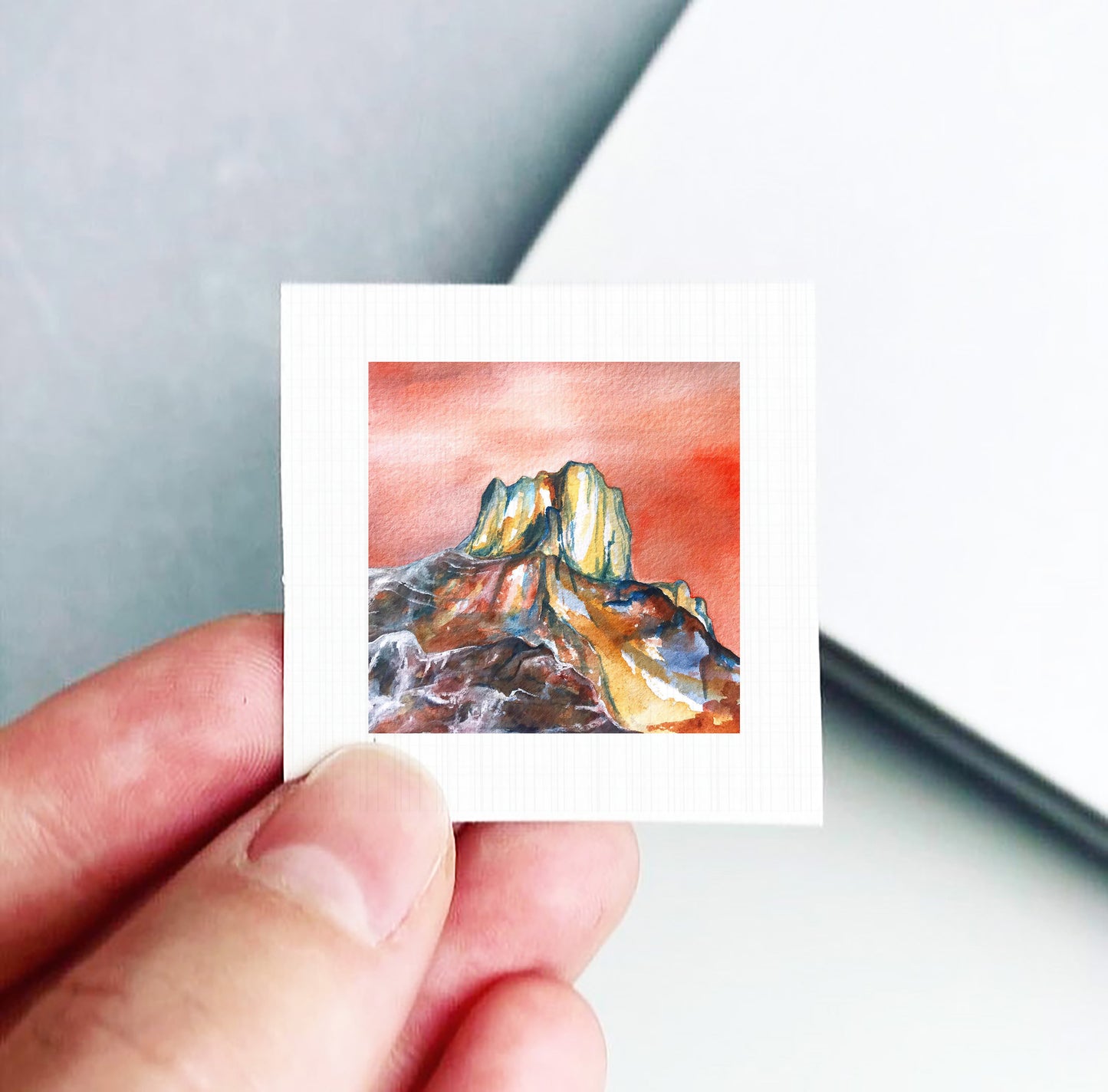 A hand is holding a small square piece of paper with a painting of a mountain landscape on it.