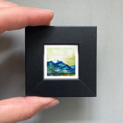 A person's hand holding a small, square-shaped black frame containing a miniature painting of a mountain landscape.