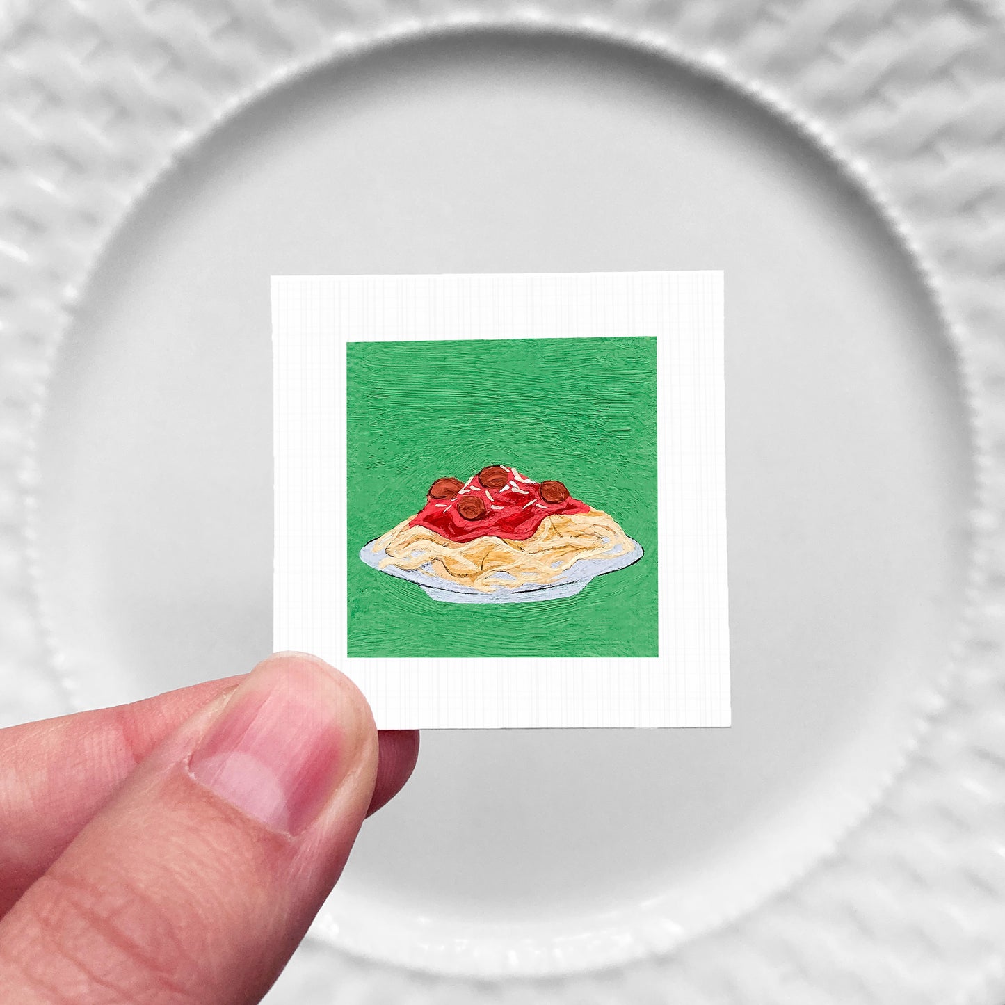 A hand is holding a small square card with an illustration of a plate of spaghetti and meatballs on it.
