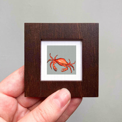 A hand is holding a small wooden frame containing a framed illustration of a crab.
