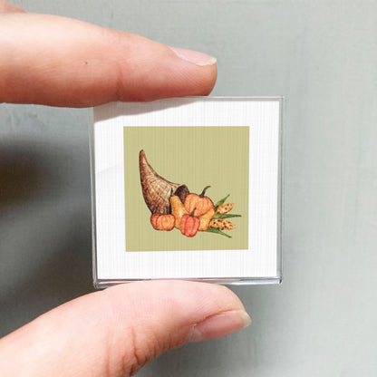 A person's hand holding a small square with a picture of a Thanksgiving-themed illustration, including a cornucopia filled with various fall fruits and vegetables.