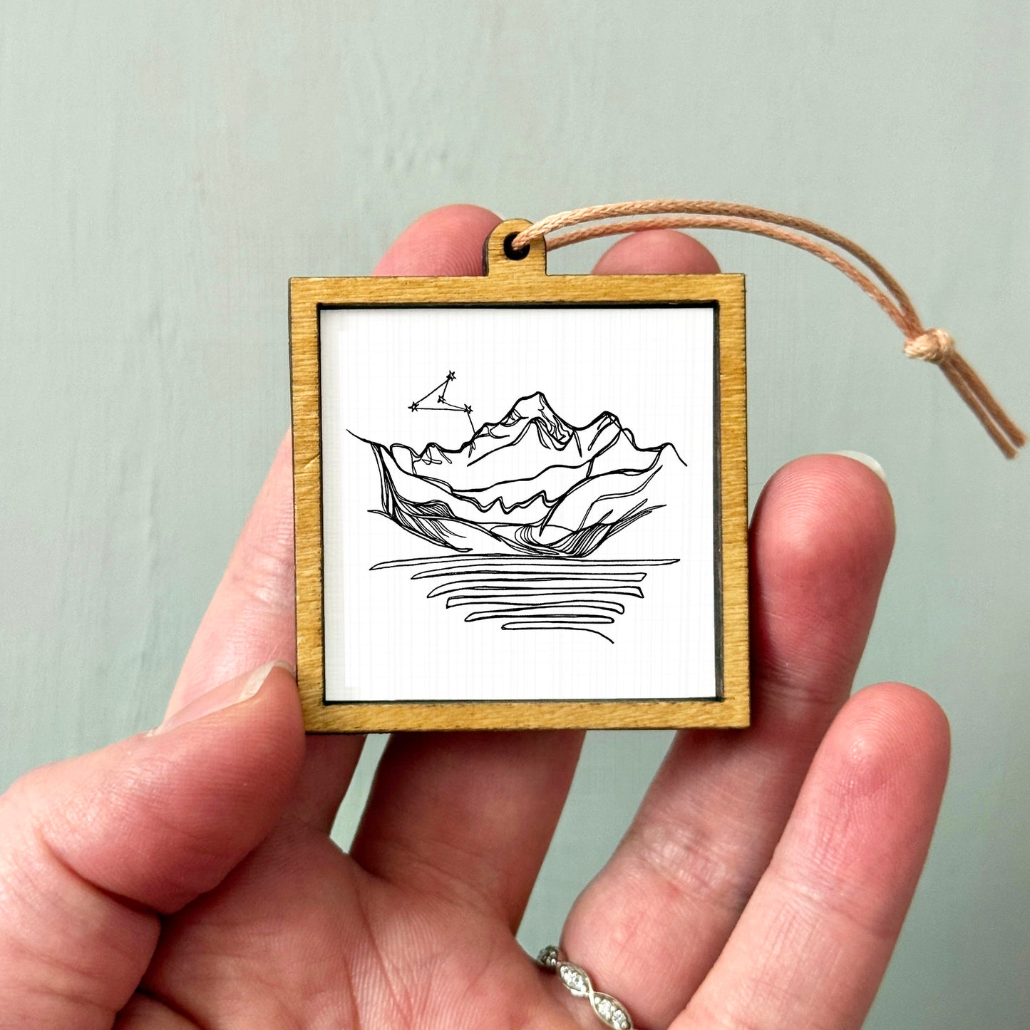 A hand holding a small wooden frame containing a black and white line drawing of a mountain landscape.