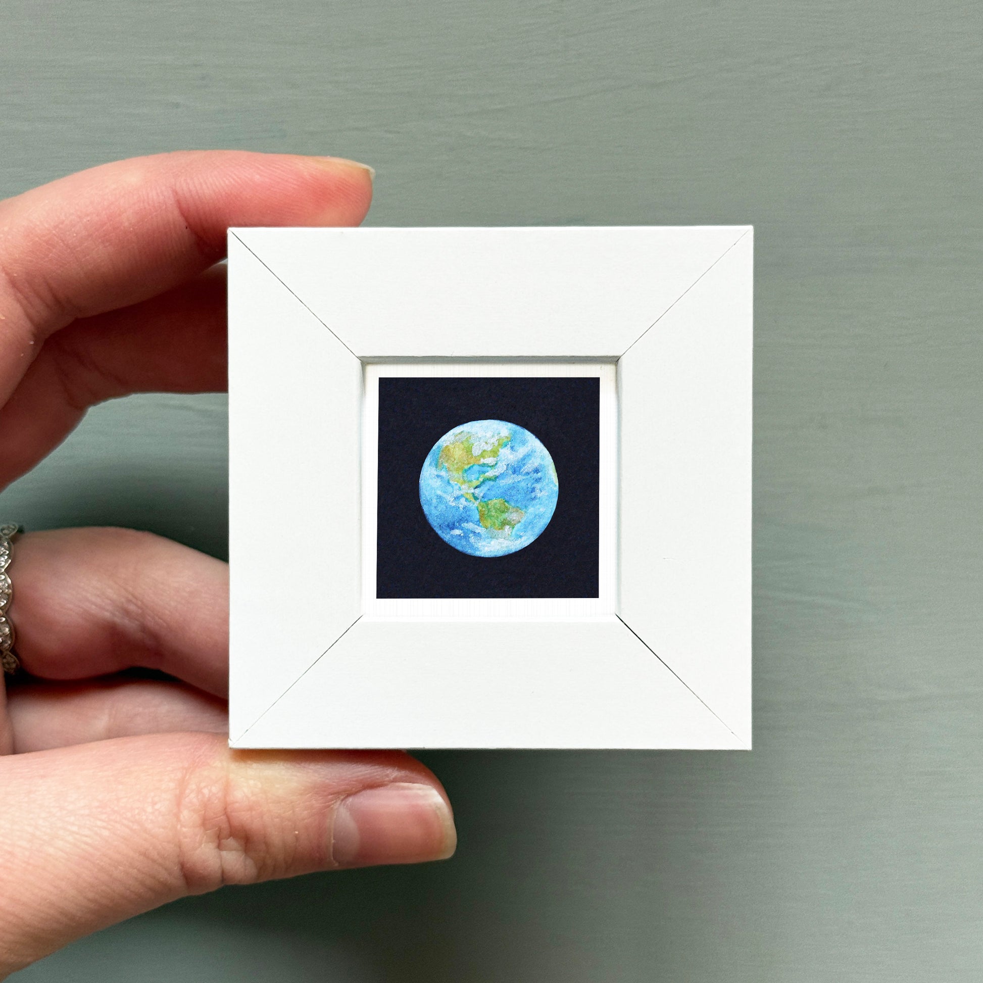 A hand is holding a small white picture frame containing a miniature representation of the Earth.