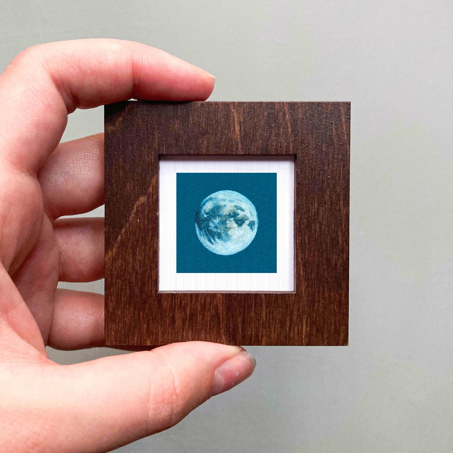 A hand is holding a small wooden frame containing a framed image of a full moon.