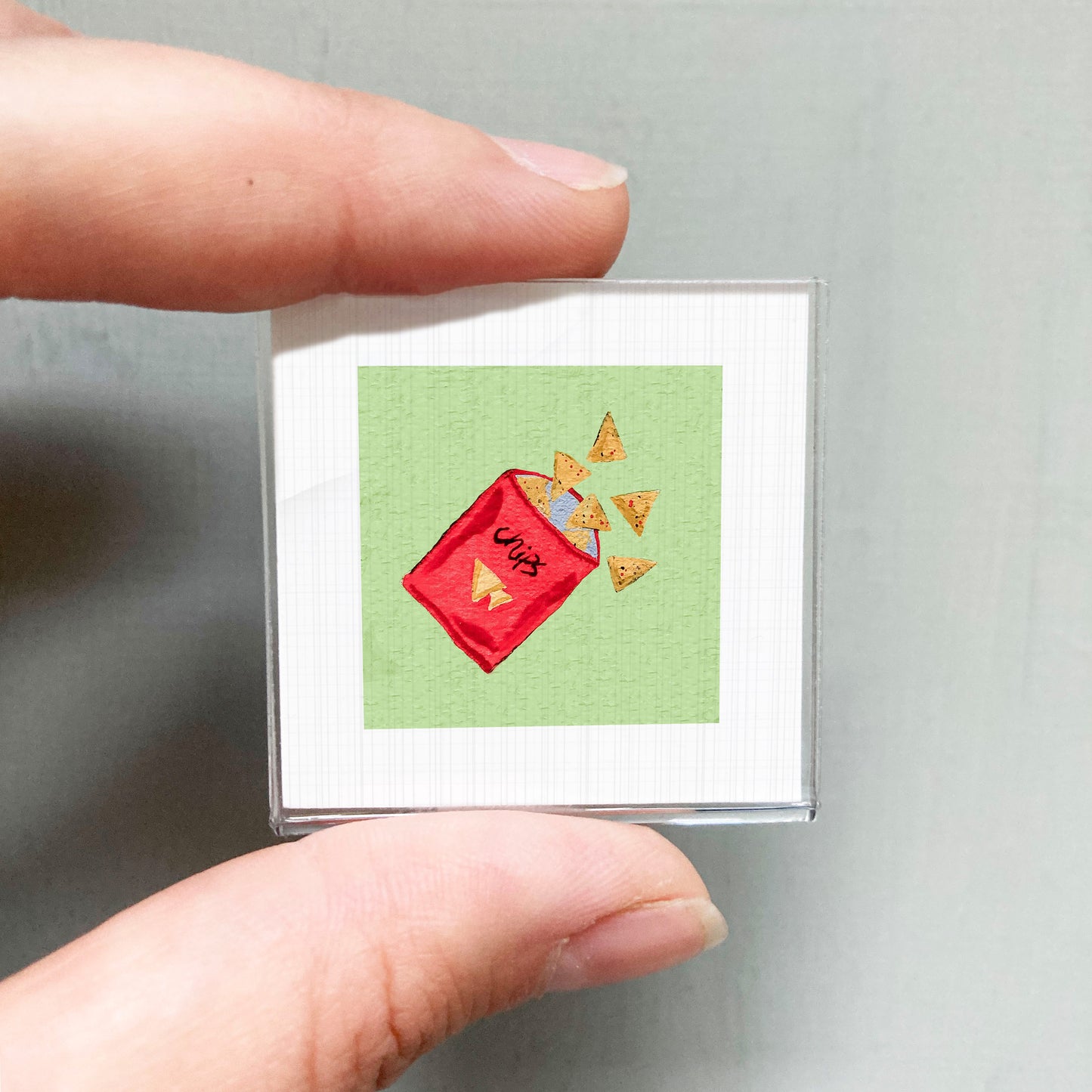 A person's hand holding a small square with a red bag of chips spilling out onto a green background.
