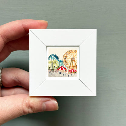 A hand is holding a small white picture frame containing an illustration of a carnival scene with ferris wheels and other rides.