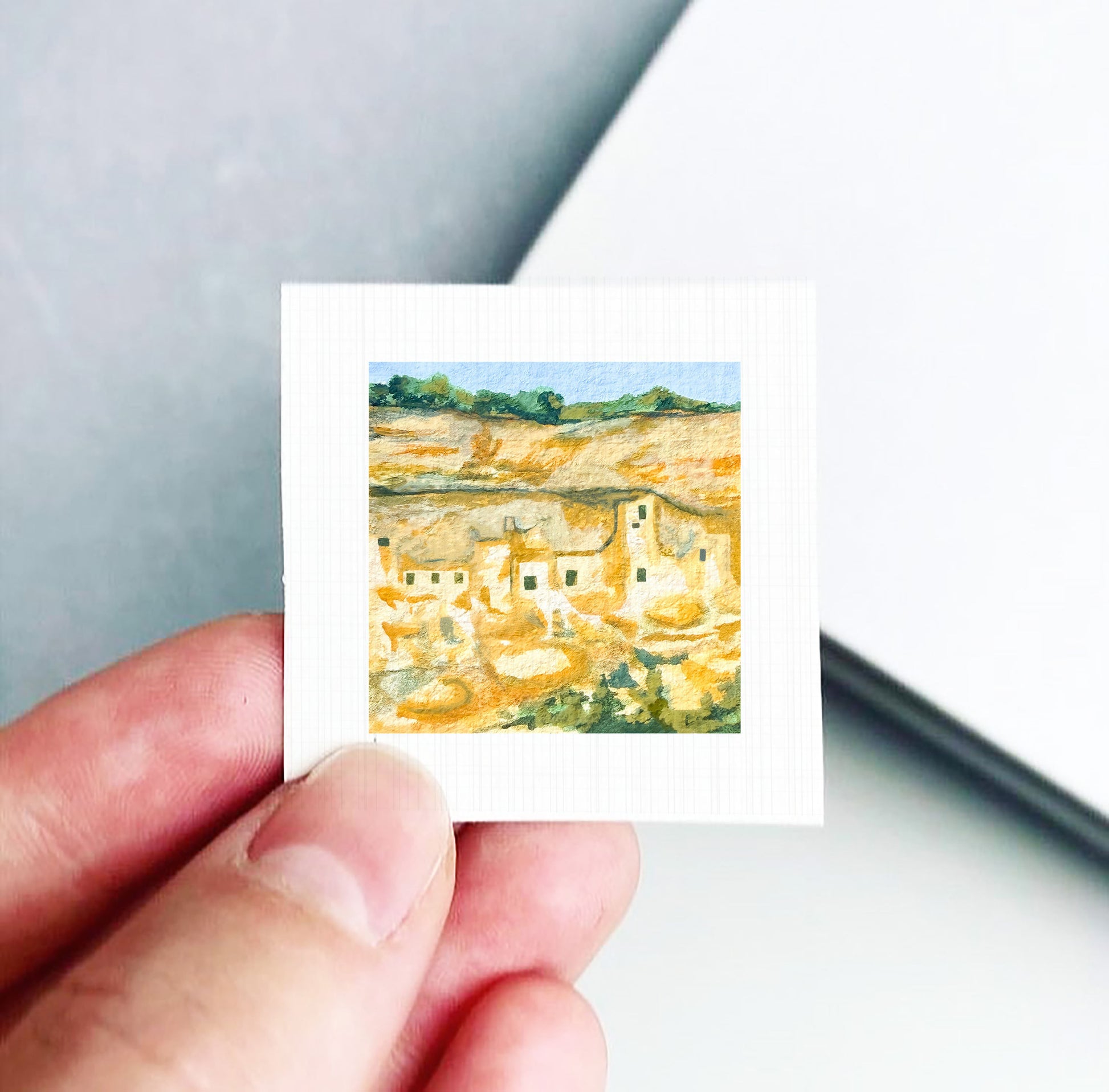 A hand is holding a small square-shaped piece of paper with a painting of a desert landscape on it.