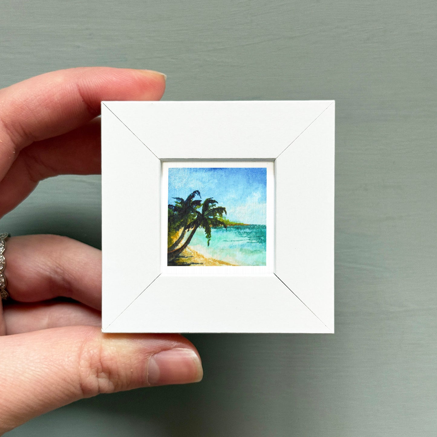A hand is holding a small white square frame containing a painting of a tropical beach scene with palm trees and a blue sky.