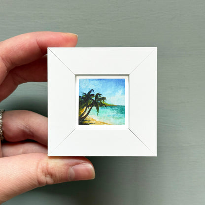 A hand is holding a small white square frame containing a painting of a tropical beach scene with palm trees and a blue sky.