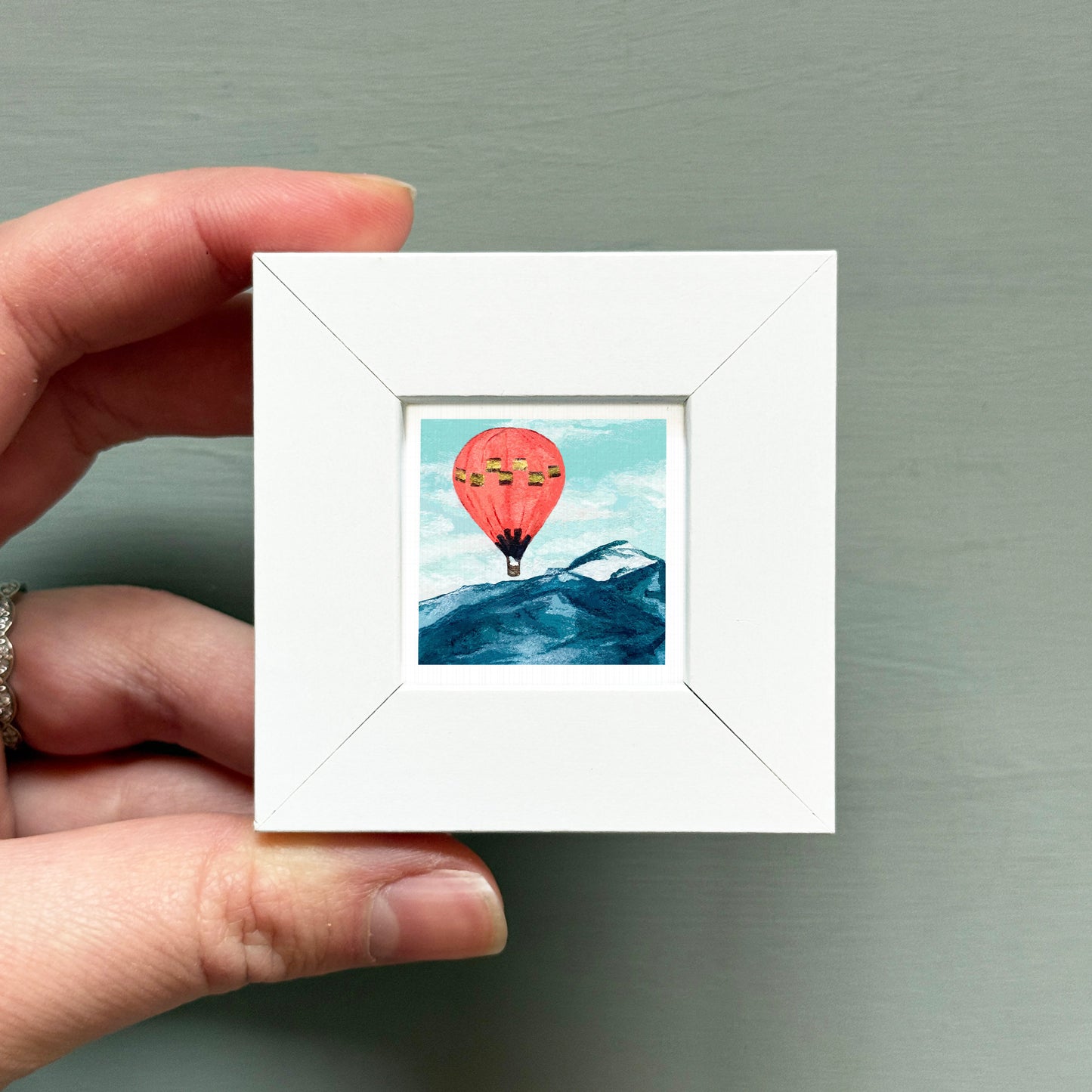A hand is holding a small white picture frame containing a painting of a hot air balloon over a mountain landscape.