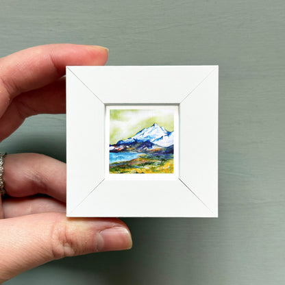 A hand is holding a small white square frame containing a watercolor painting of a mountain landscape.