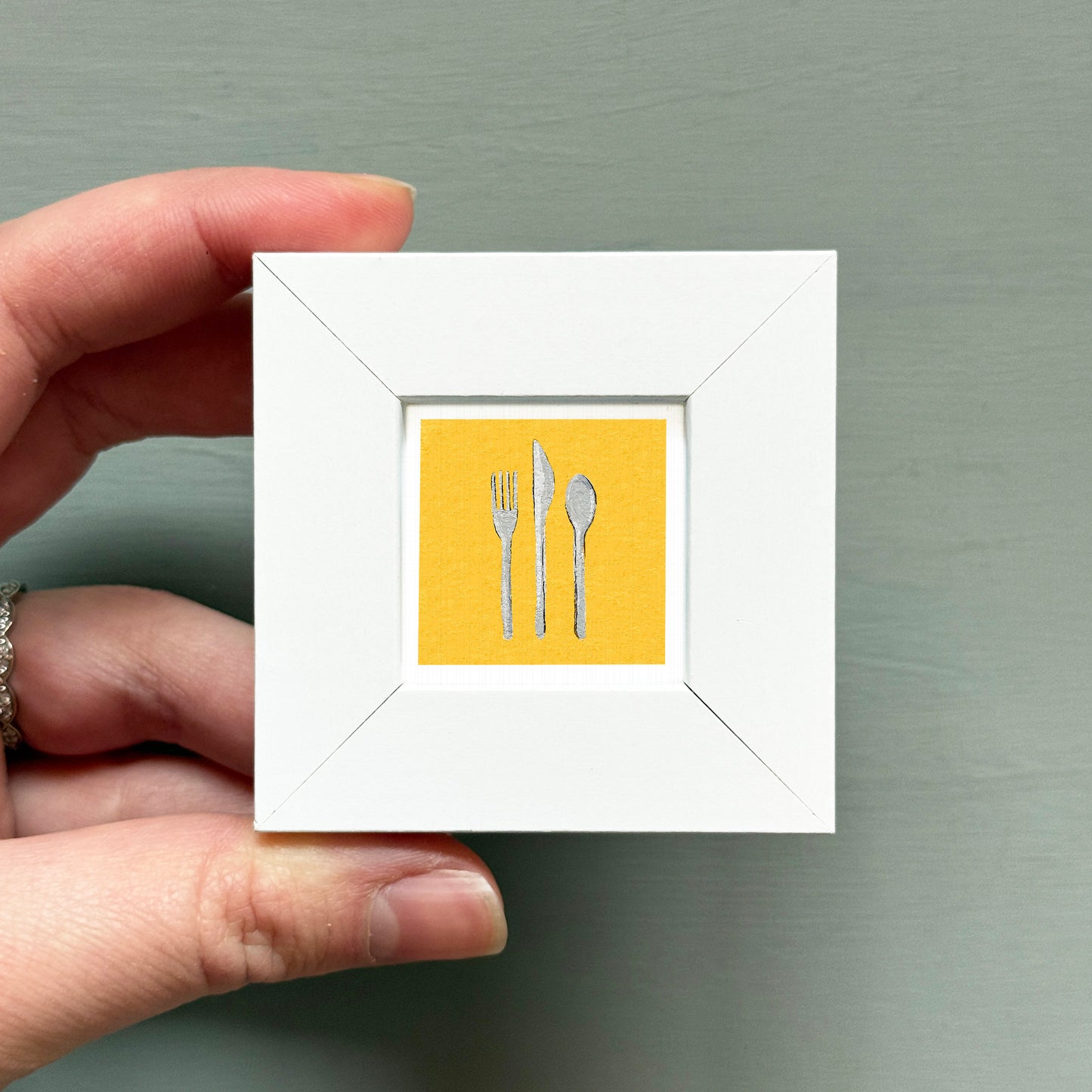 A hand is holding a small white square frame containing a yellow square with a picture of a fork, knife, and spoon.