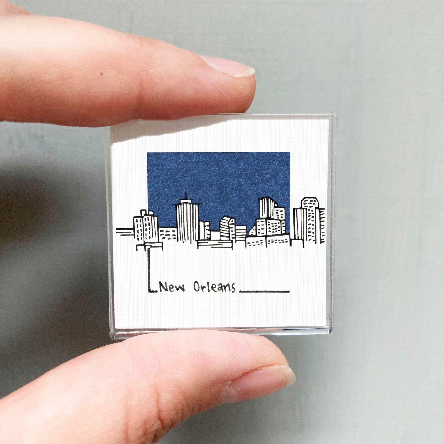 A person's hand holding a small square magnet with a drawing of a city skyline and the text "New Orleans" written below it.