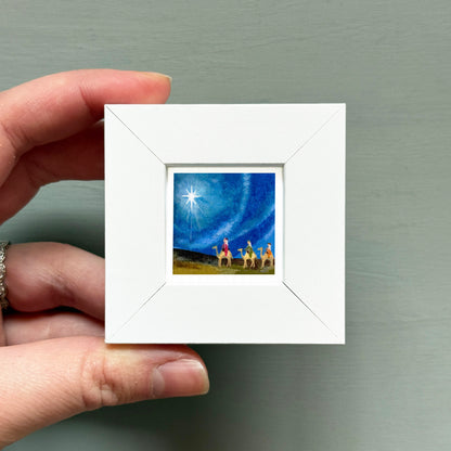 A hand is holding a small white square frame containing a painting of a starry night sky with a group of people on camels.