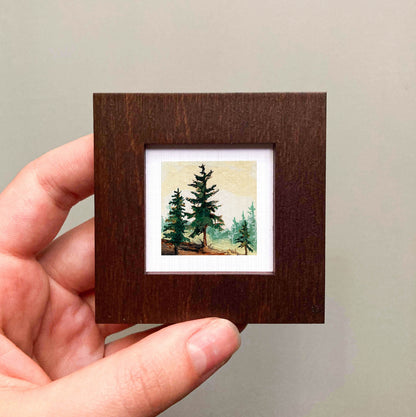 A hand is holding a small wooden frame containing a painting of a forest scene with pine trees.