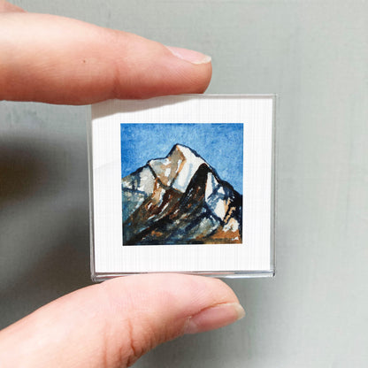 A person's hand holding a small square picture of a mountain landscape.