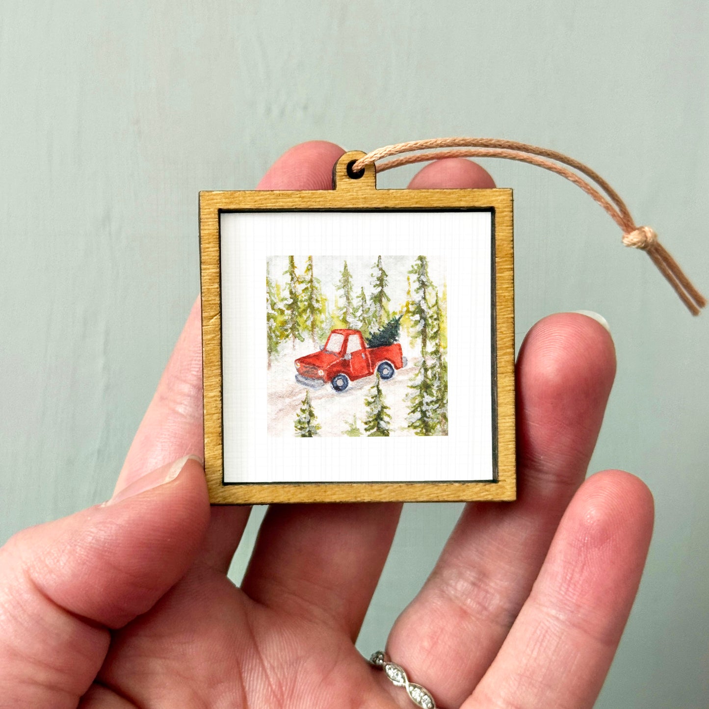 A hand holding a small wooden frame containing a picture of a red truck driving through a snowy forest.