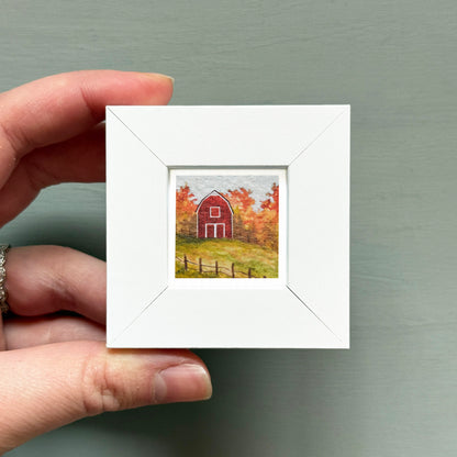A hand is holding a small white picture frame containing a painting of a red barn surrounded by trees in autumn colors.