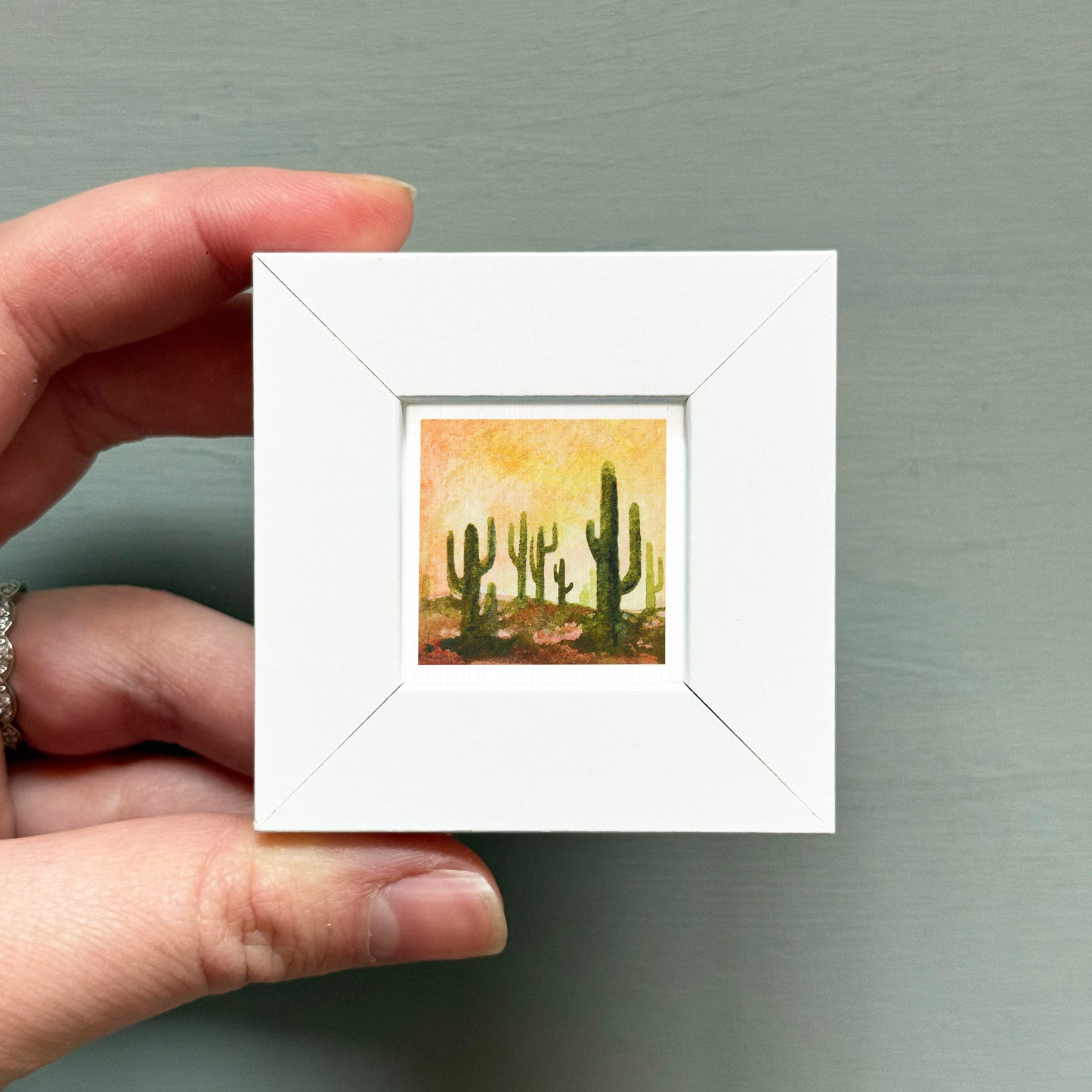 A hand is holding a small white square frame containing a painting of cacti.
