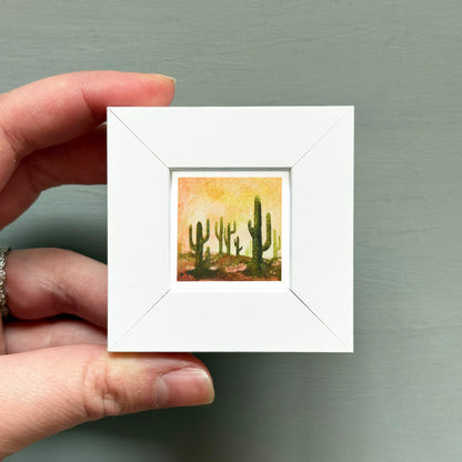 A hand is holding a small white square frame containing a painting of cacti.