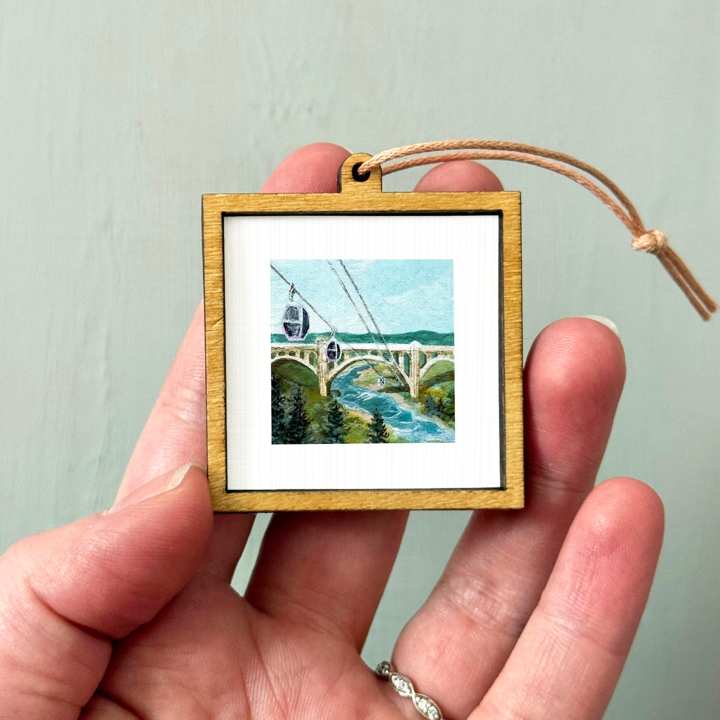 A person's hand holding a small wooden frame containing a miniature painting of a bridge and river.