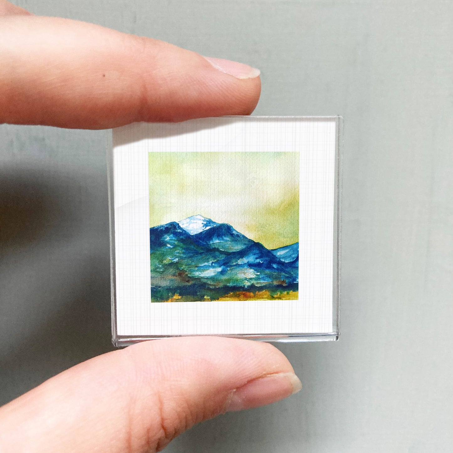 A person's hand holding a small square picture of a mountain landscape.