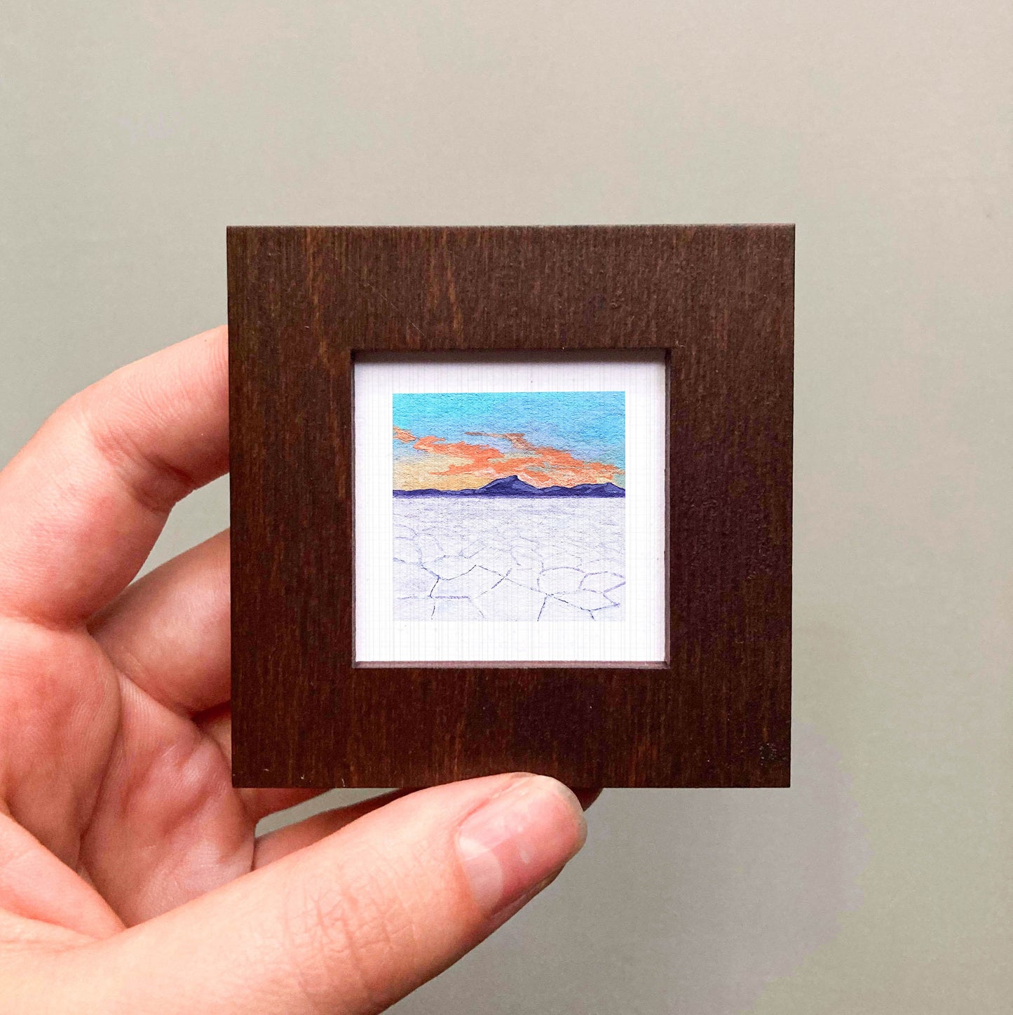 A hand is holding a small, square-shaped wooden frame containing a miniature painting of a landscape with mountains and a colorful sky.