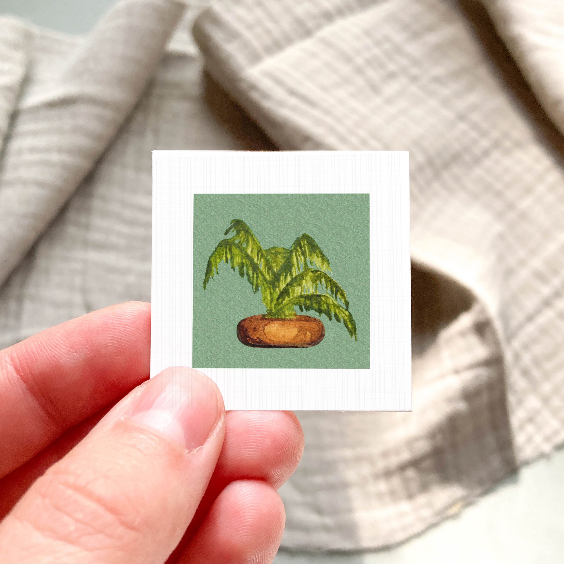 A hand is holding a small square card with a painting of a palm tree on it.