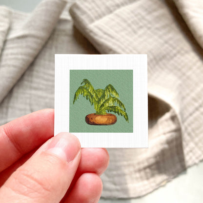 A hand is holding a small square card with a painting of a palm tree on it.