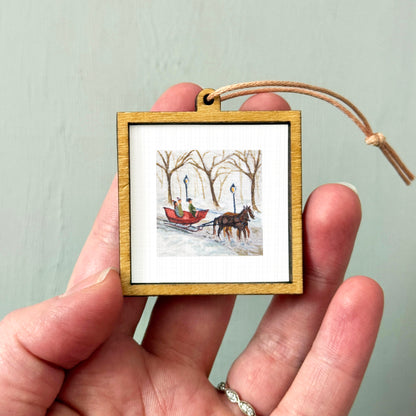 A person's hand is holding a small wooden frame containing a framed painting of a horse-drawn sleigh in a snowy landscape.