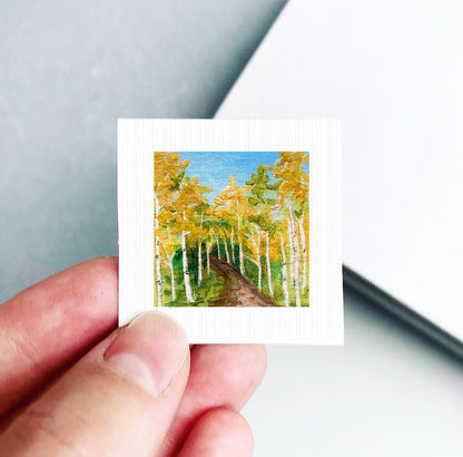 A hand holding a small square picture of a forest with yellow trees against a blue sky.