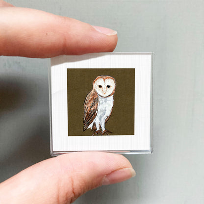 A person's hand is holding a small square picture of an owl against a brown background.