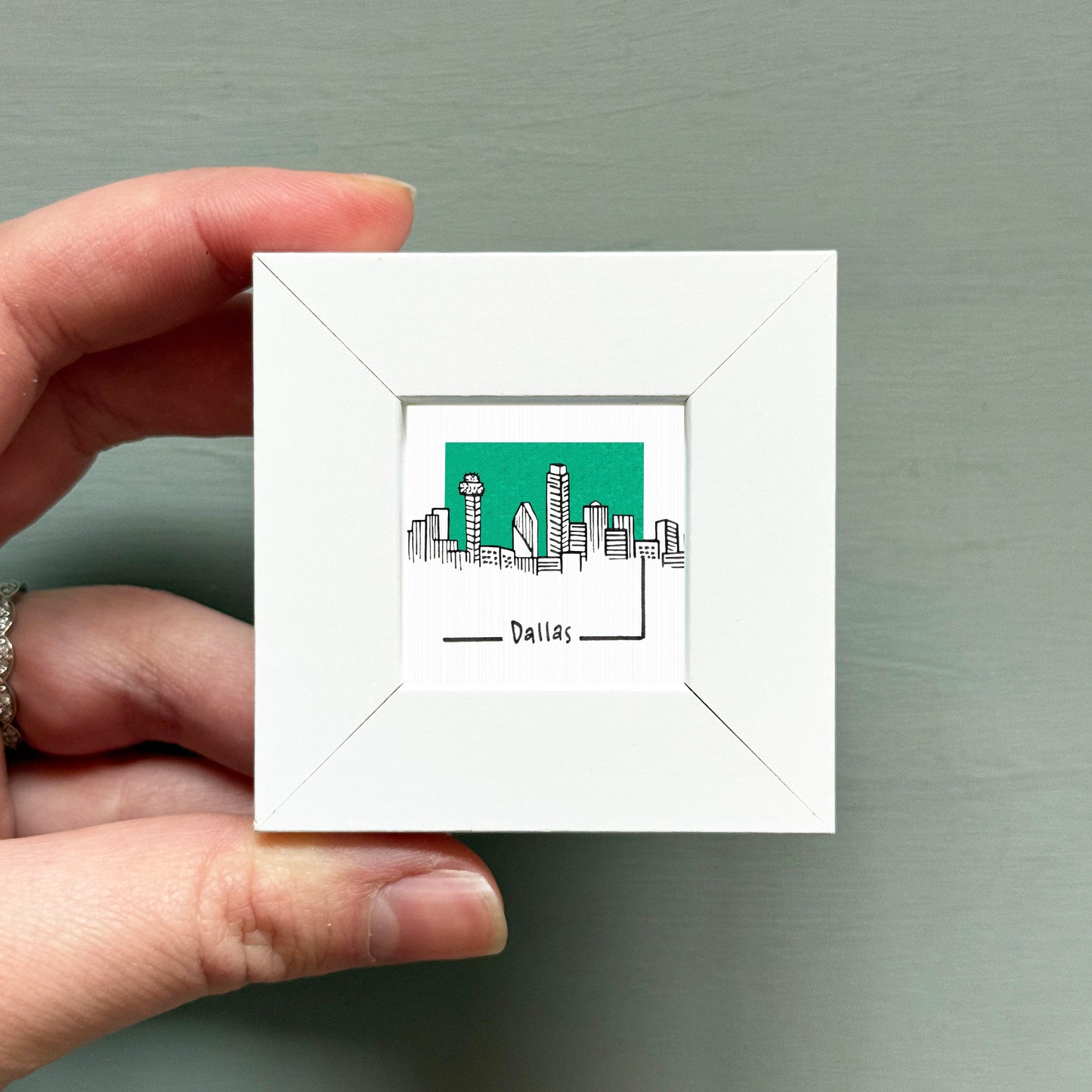 A hand is holding a small white square frame containing a drawing of the skyline of Dallas, Texas.