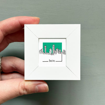 A hand is holding a small white square frame containing a drawing of the skyline of Dallas, Texas.