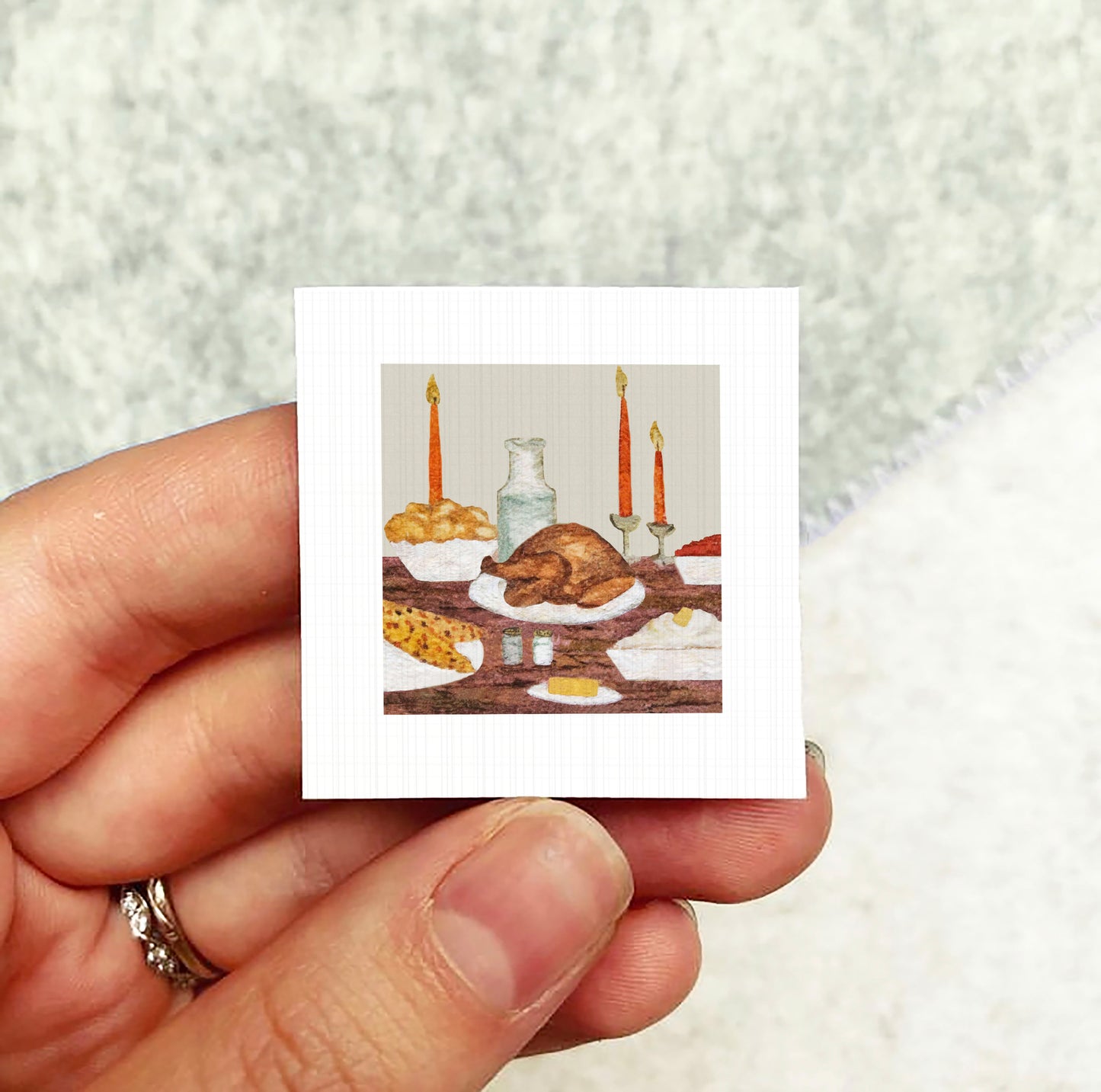 A hand holding a small square picture depicting a table with a roasted turkey, bread, and candles.