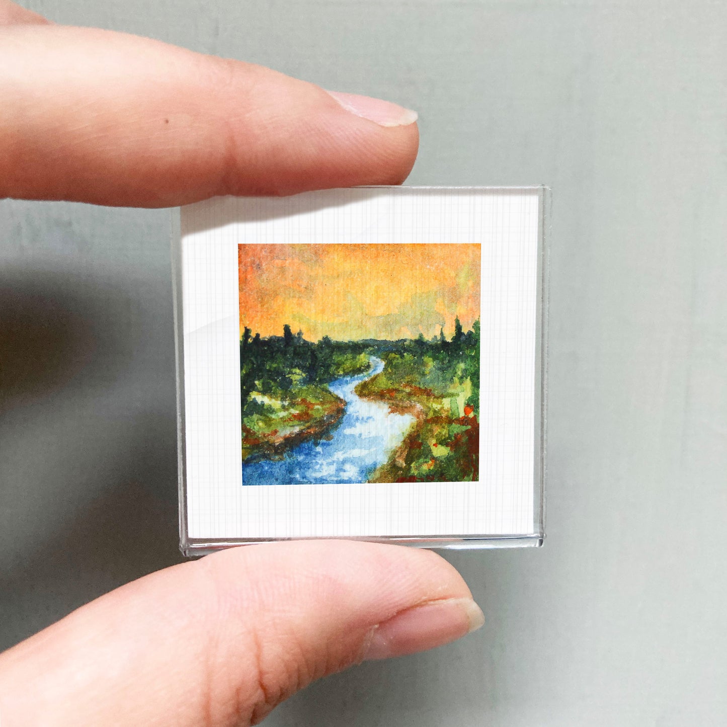 A person's hand holding a small square picture of a landscape with a river and trees.