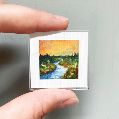 A person's hand holding a small square picture of a landscape with a river and trees.