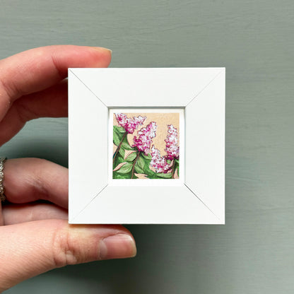 A hand is holding a small white square frame containing a painting of pink flowers.