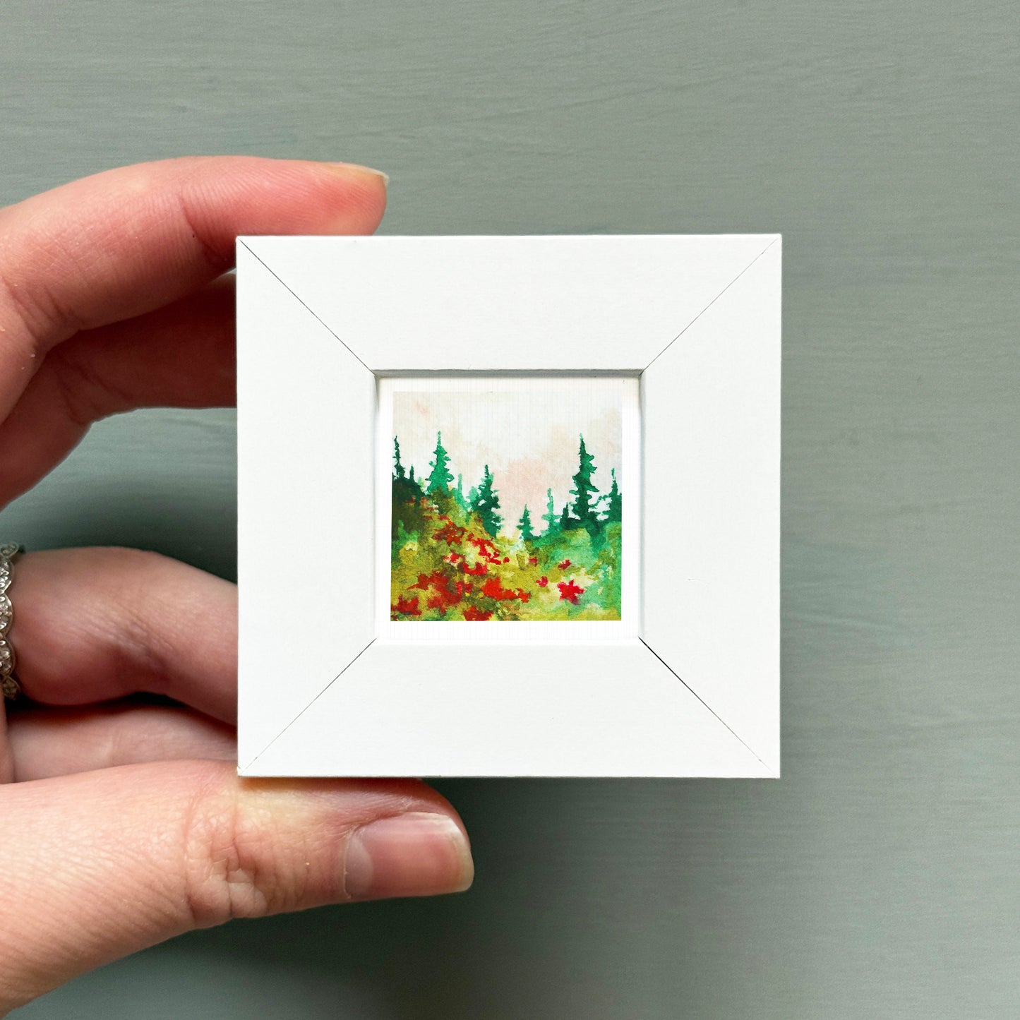 A hand is holding a small white square frame containing a painting of a forest scene with green trees and red flowers.