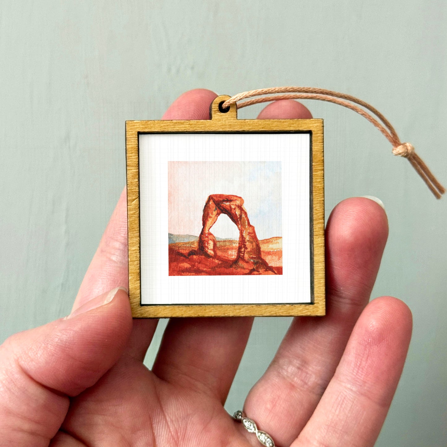 A person's hand holding a small wooden frame containing a framed painting of a natural arch formation.