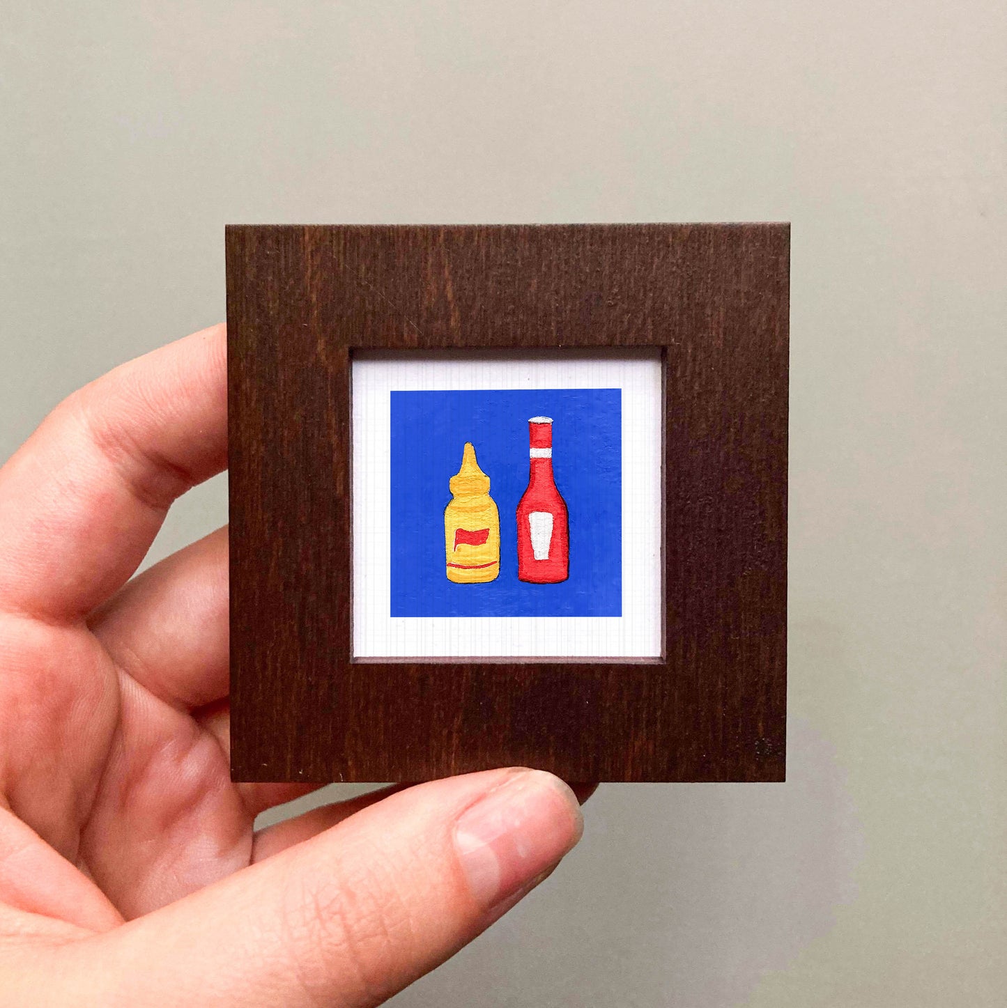 A hand is holding a small square frame containing two bottles of condiments, one yellow and one red, against a blue background.