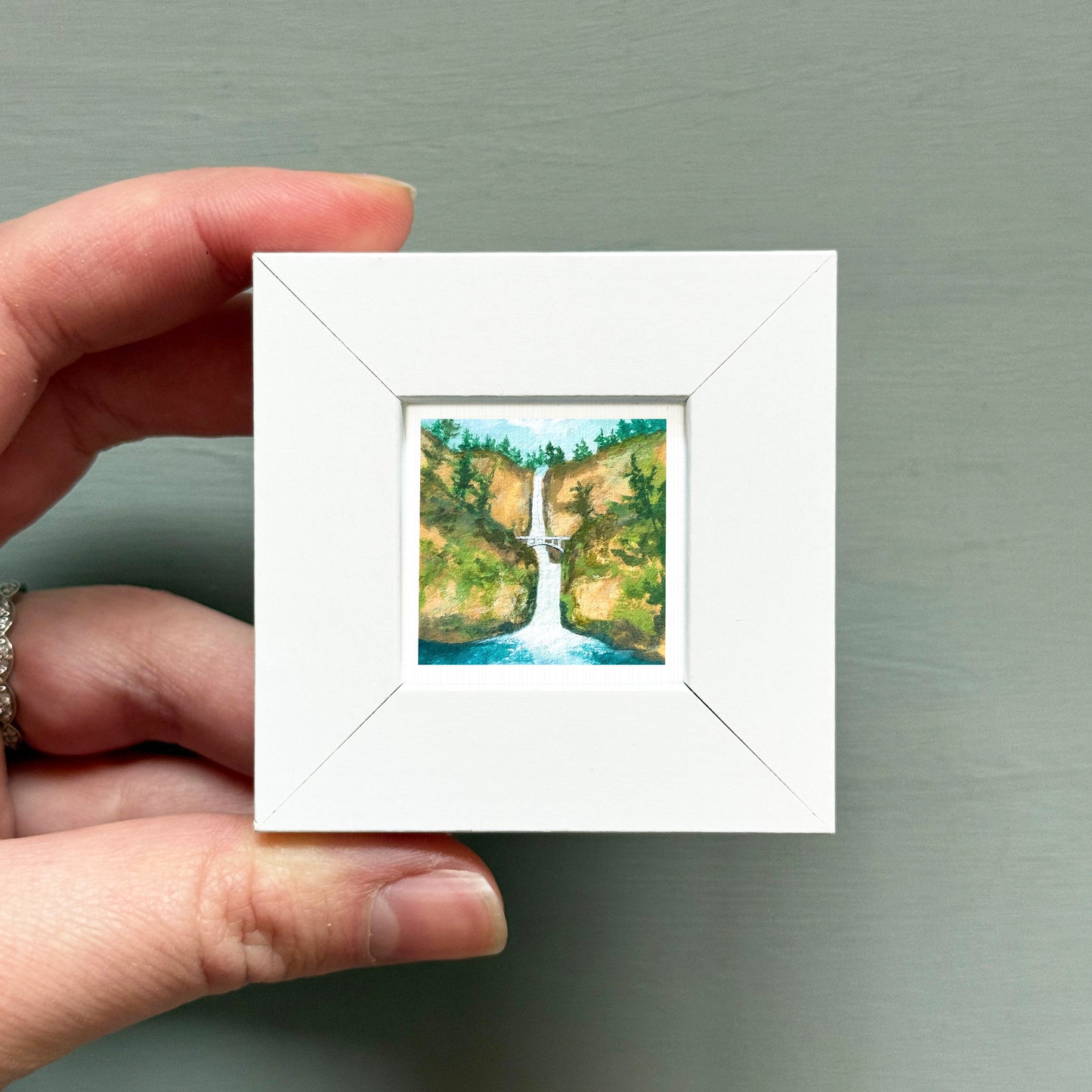 A hand is holding a small white square frame containing a painting of a waterfall surrounded by lush greenery.