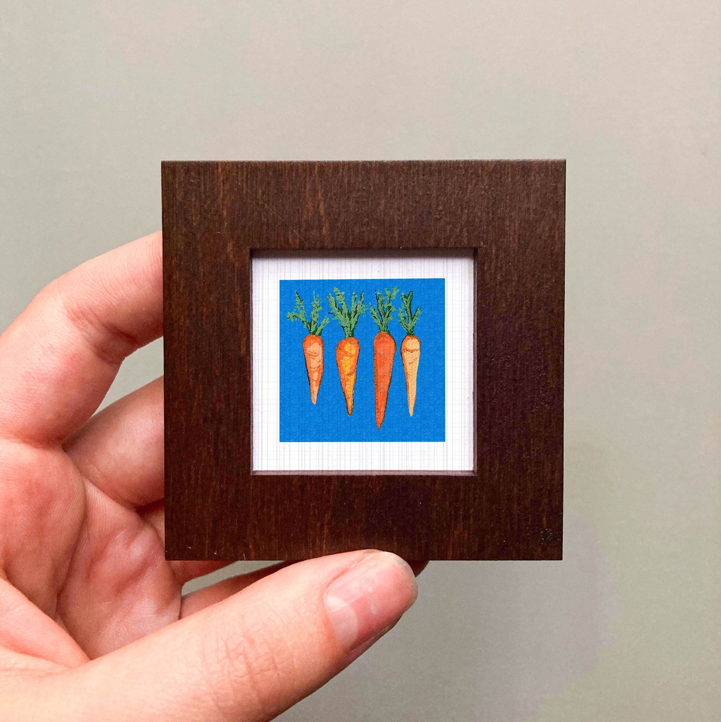 A hand is holding a small square frame containing a painting of three carrots against a blue background.