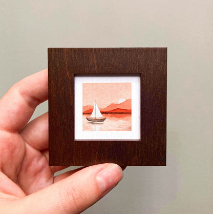 A hand is holding a small wooden frame containing a painting of a sailboat on a body of water.