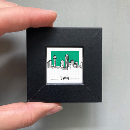 A hand is holding a small black square frame containing a miniature illustration of the Dallas skyline.